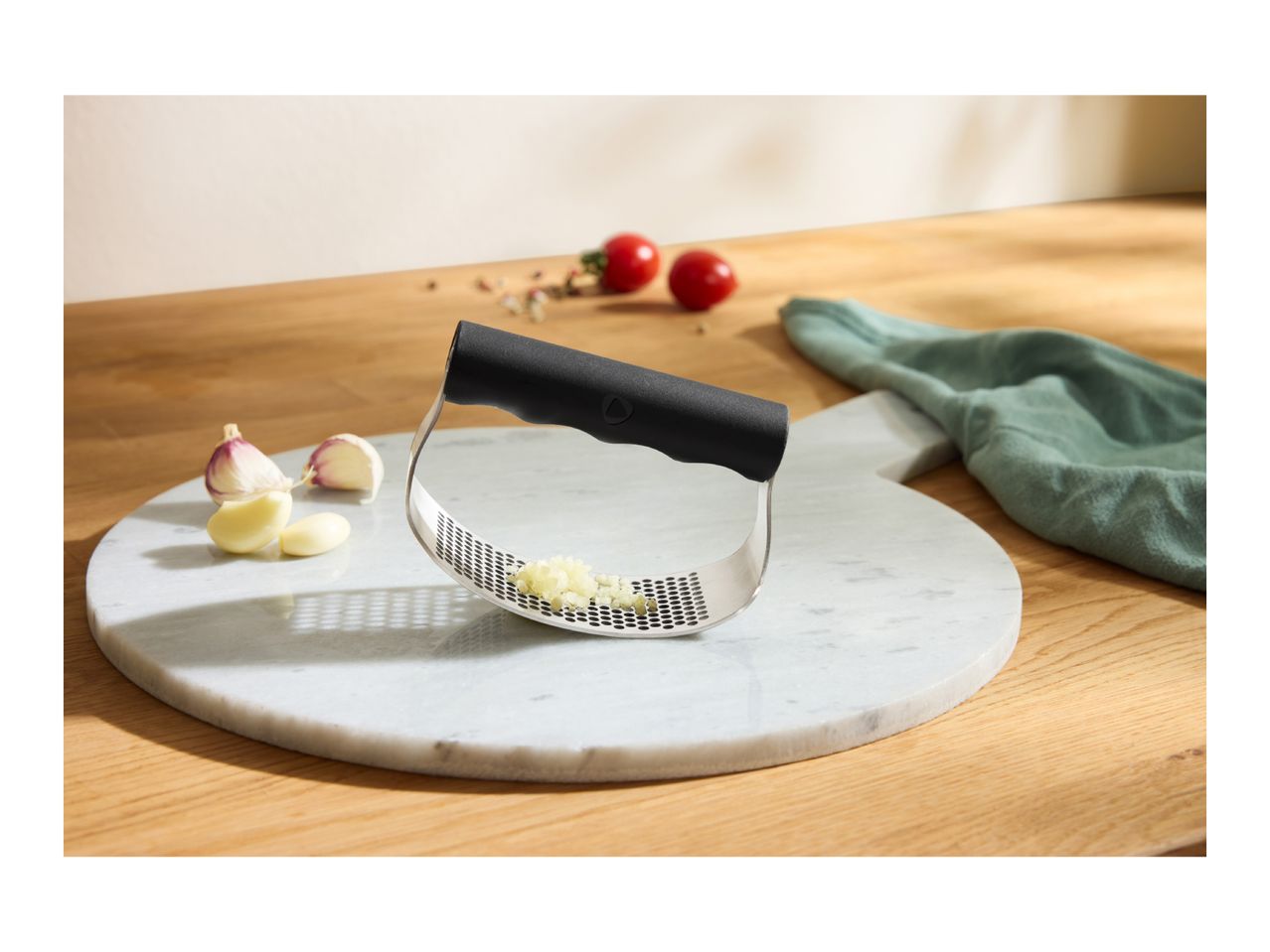 Garlic press with crushed garlic, garlic cloves, and tomatoes on a marble board.