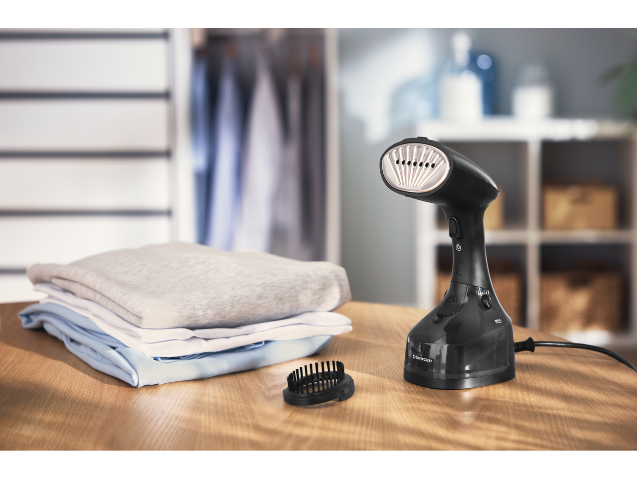 Vaporizador de roupa preto com escova e roupas dobradas numa mesa de madeira.