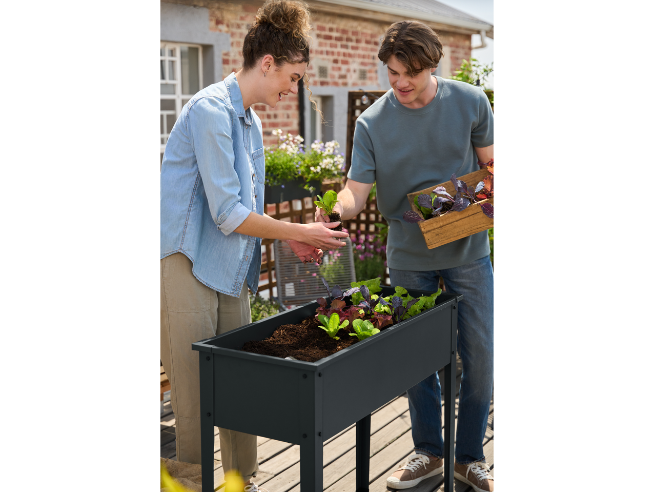 Two people planting seedlings in a raised garden bed on a sunny patio.