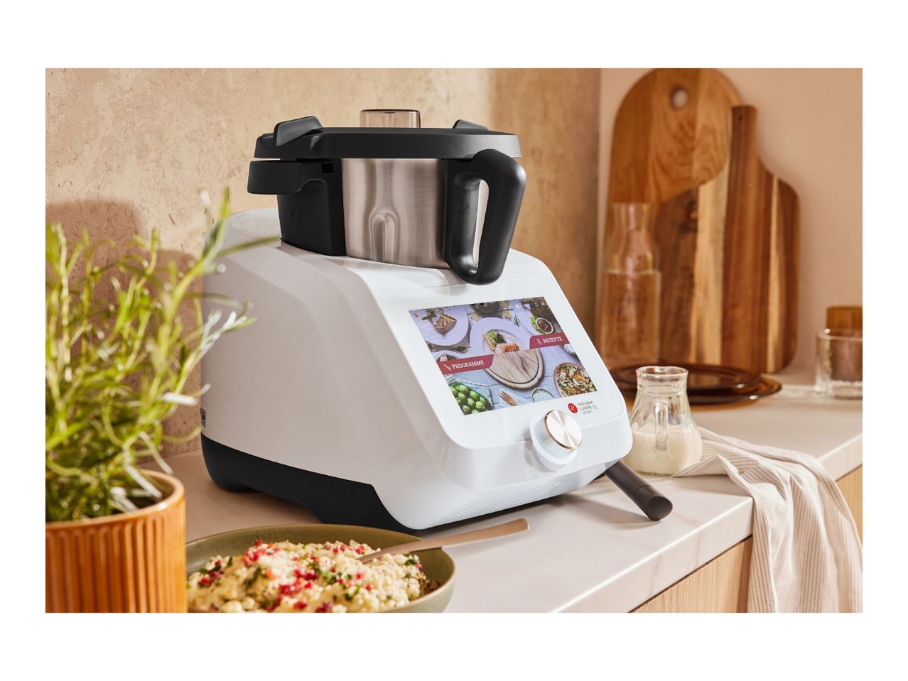 A white Monsieur Cuisine Smart food processor on a kitchen counter with a bowl of food.