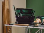 Green and black tool bag with tools, a drill, and wood planks on a table.