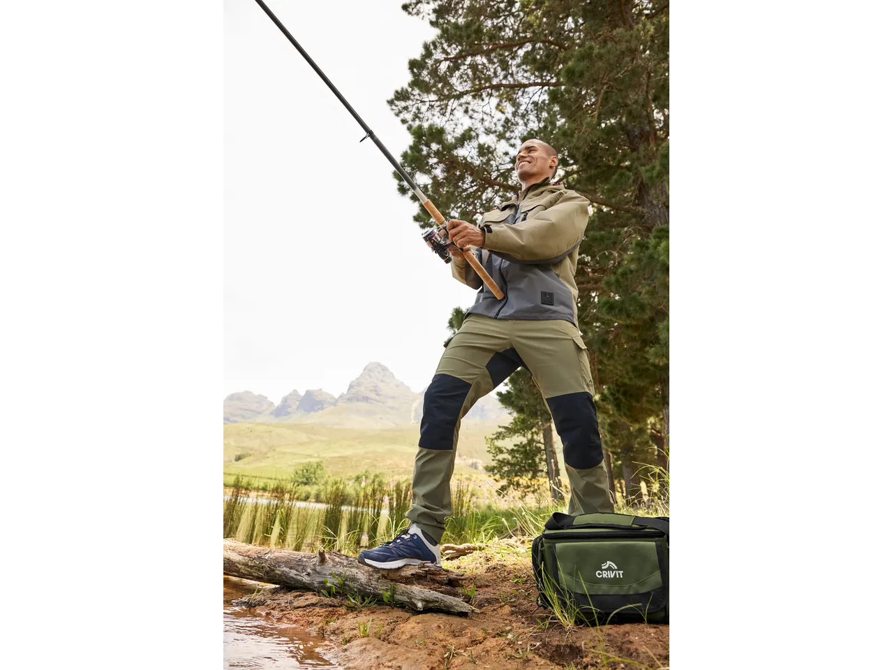 Man fishing in outdoor gear, wearing a two-tone jacket and cargo pants, with a cooler bag.