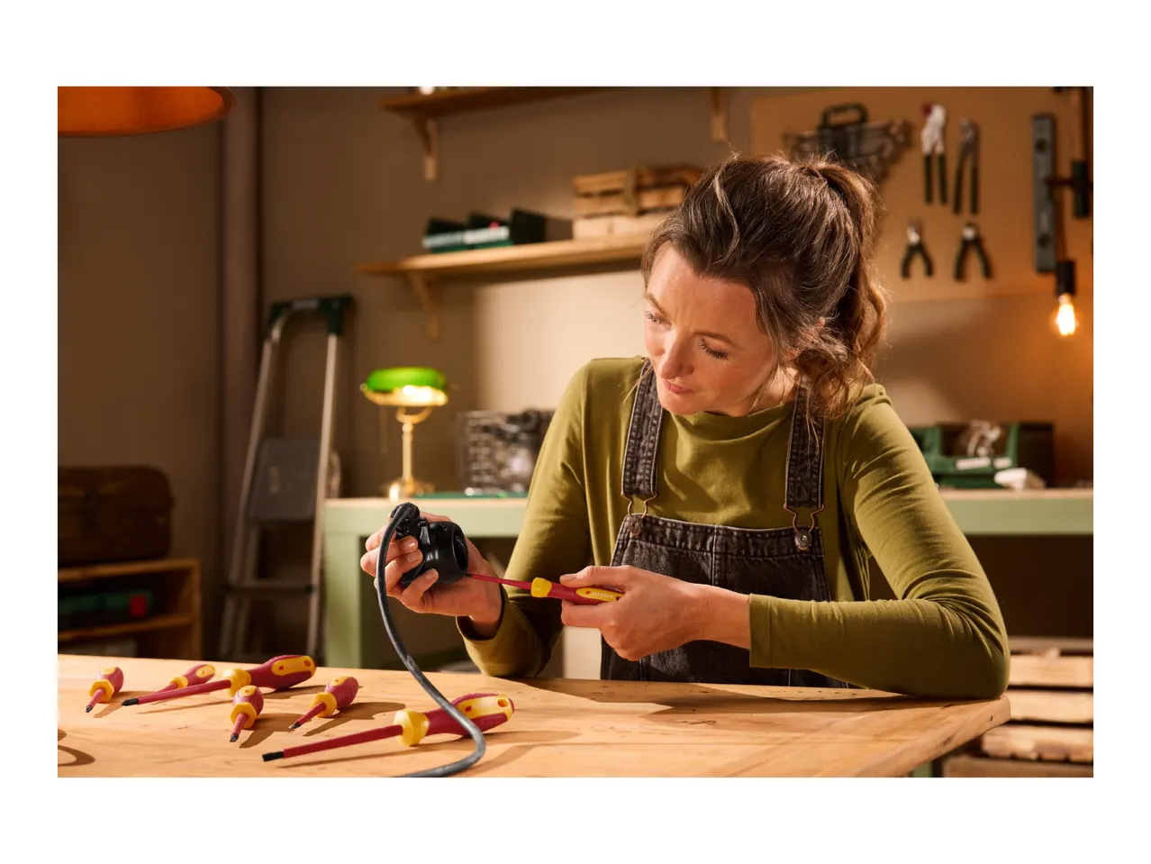 A woman in a workshop uses a screwdriver to work on an electrical component, with other screwdrivers on the wooden table.