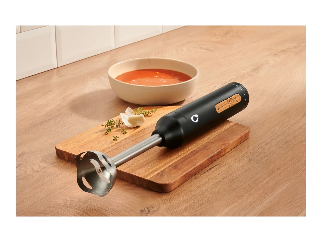 Black immersion blender on a wooden board with a bowl of tomato soup and herbs.