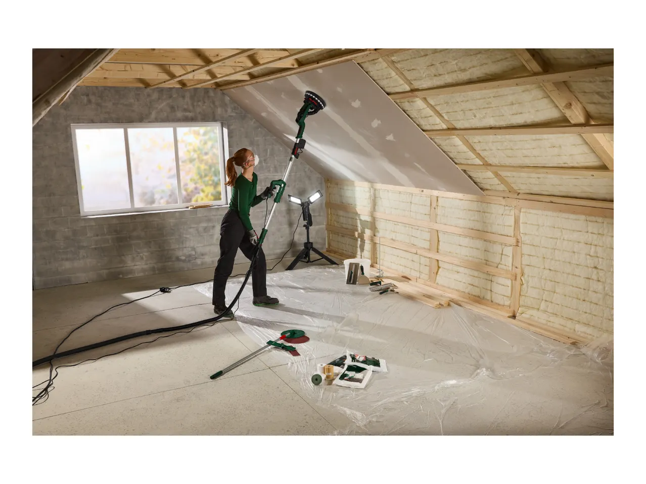 Woman sanding drywall in an attic with insulation, using a long-reach sander.
