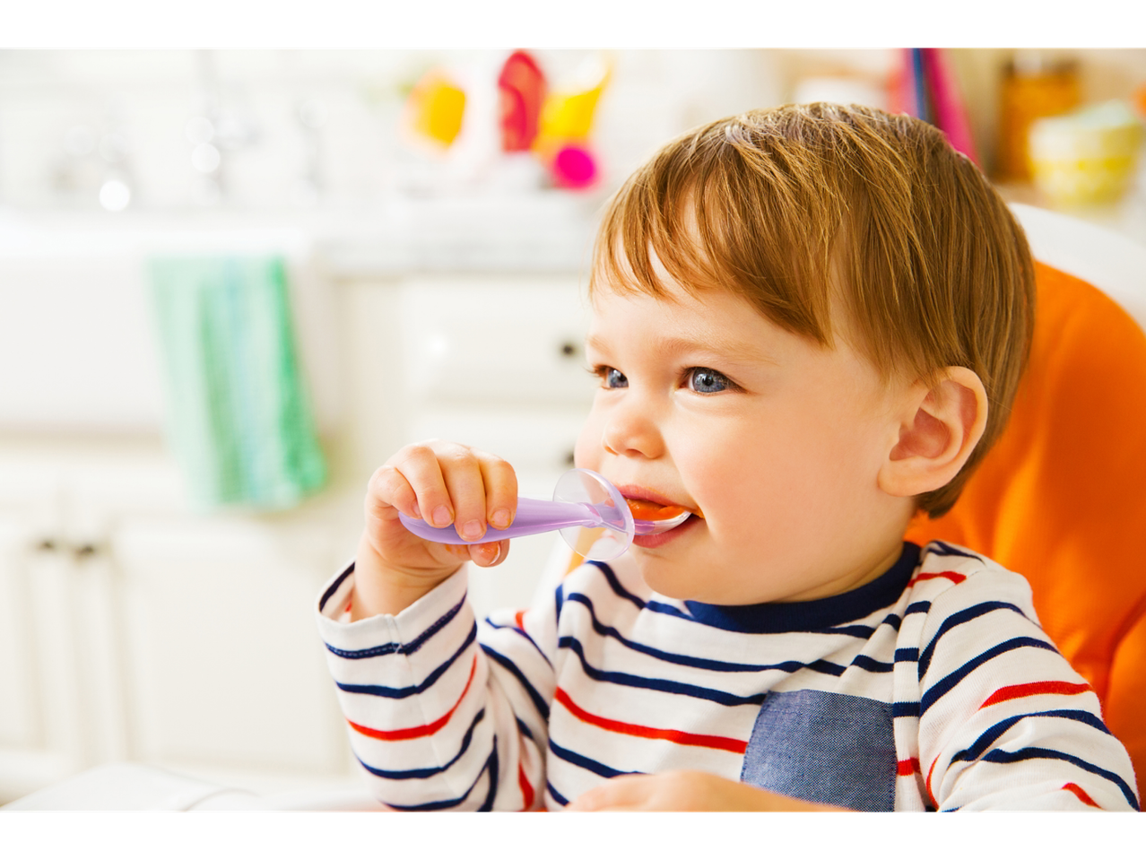 A baby in a high chair feeding himself with a purple spoon.