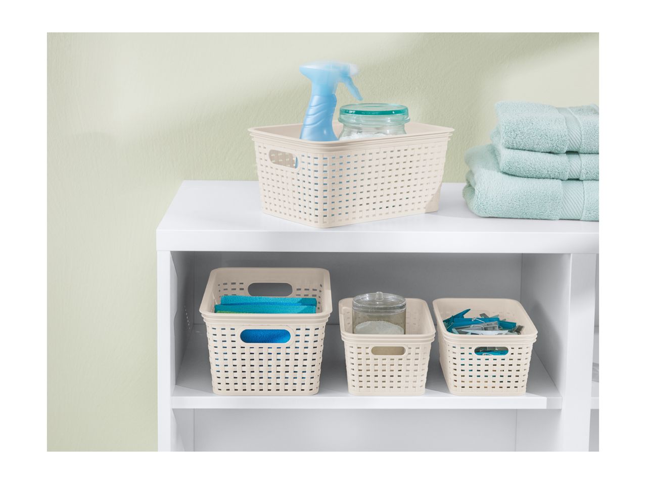 Beige plastic storage baskets with woven design hold cleaning supplies and laundry items on a white shelf.
