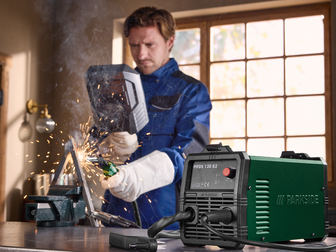 Мъж заварява метал с инверторен заваръчен апарат, искри и дим се носят във въздуха.