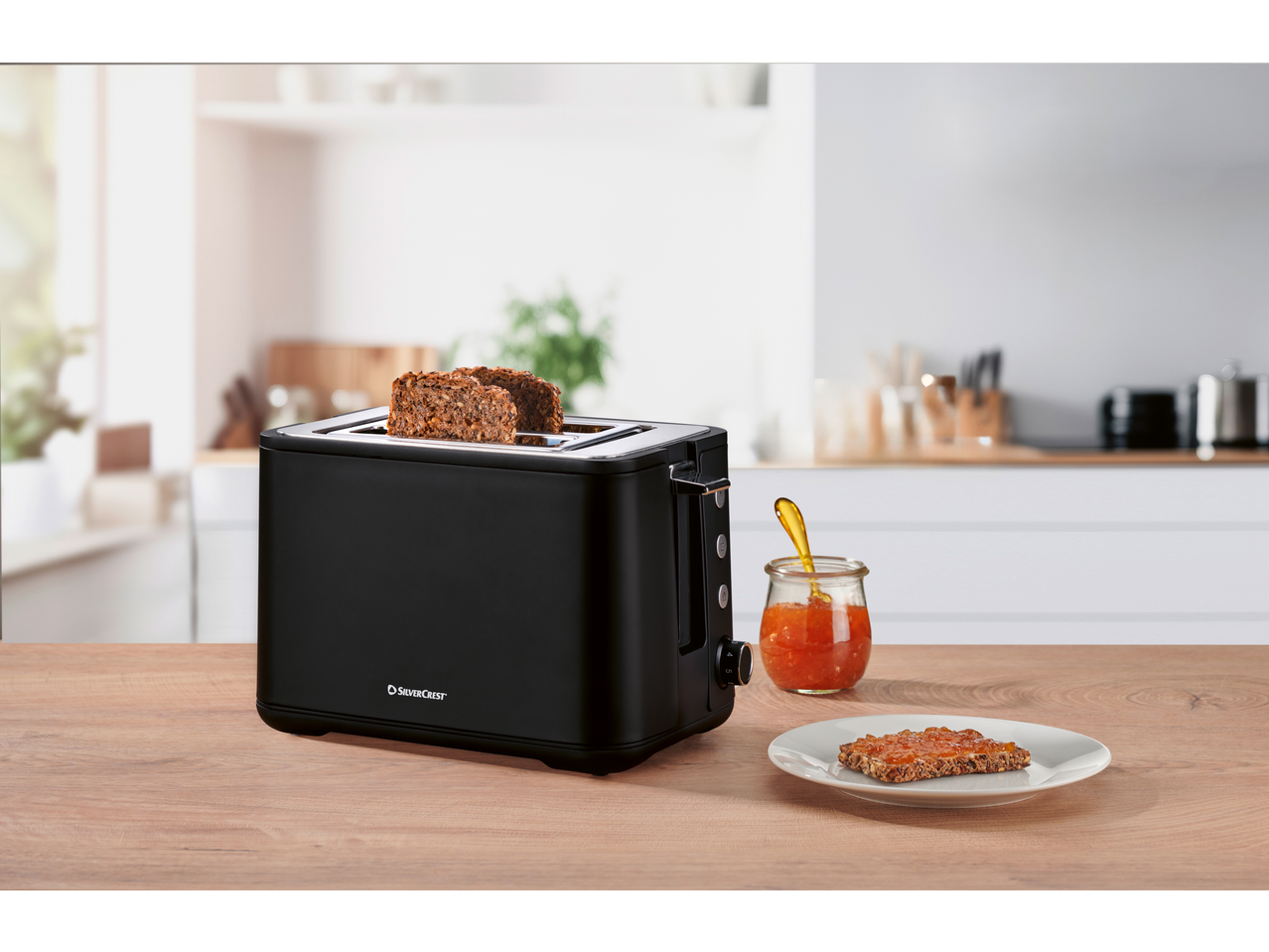 Black toaster with two slices of toast, a jar of jam, and a jam-covered toast on a plate.