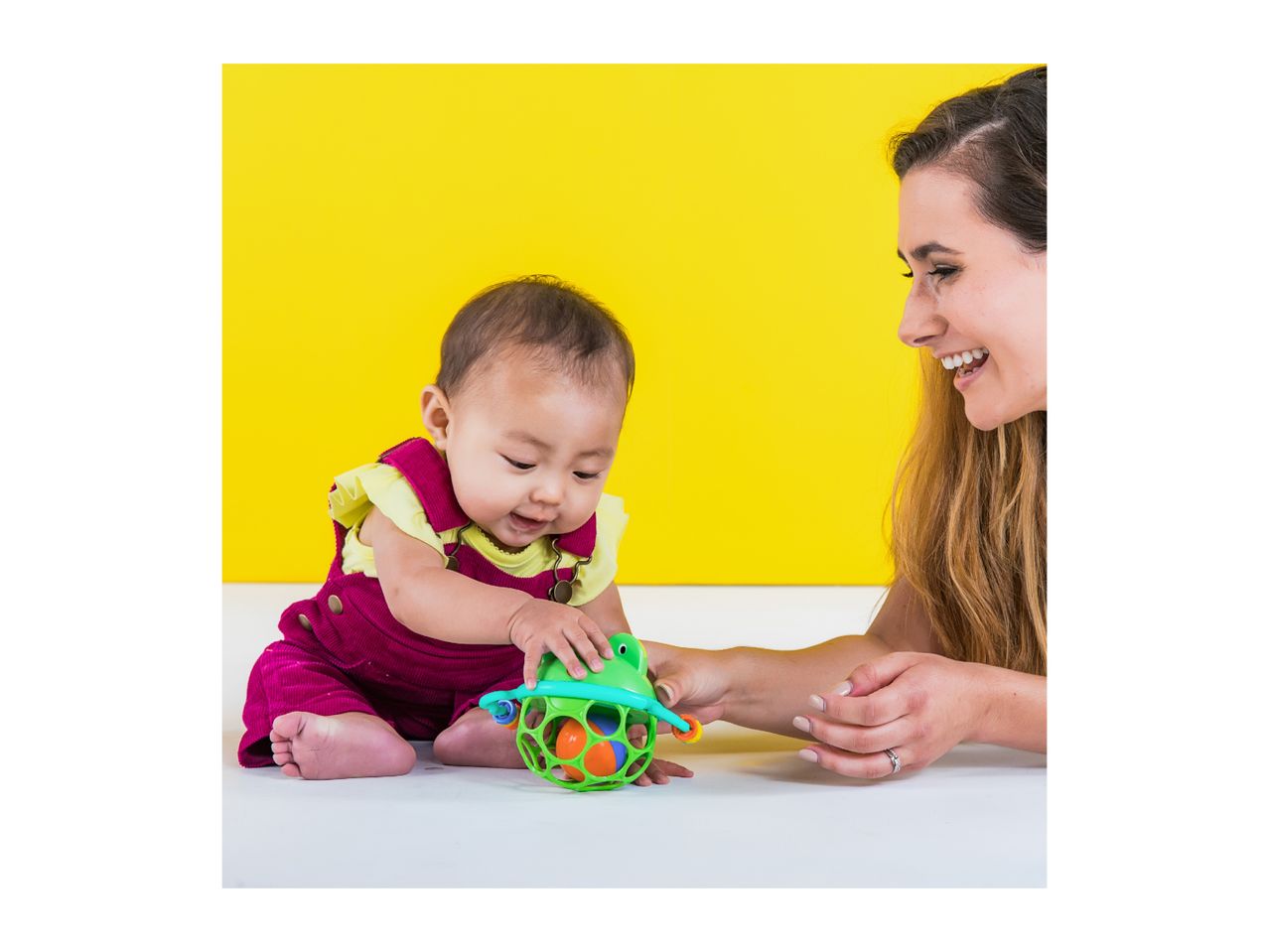 Baby and adult playing with a colorful frog-shaped rattle toy on a yellow background.