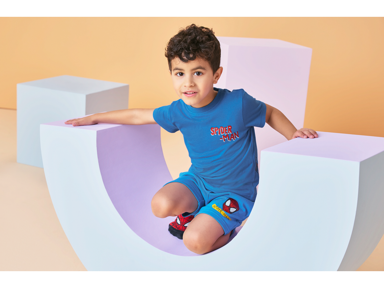 Boy in blue Spider-Man jersey shorts and matching t-shirt, sitting in a curved prop.