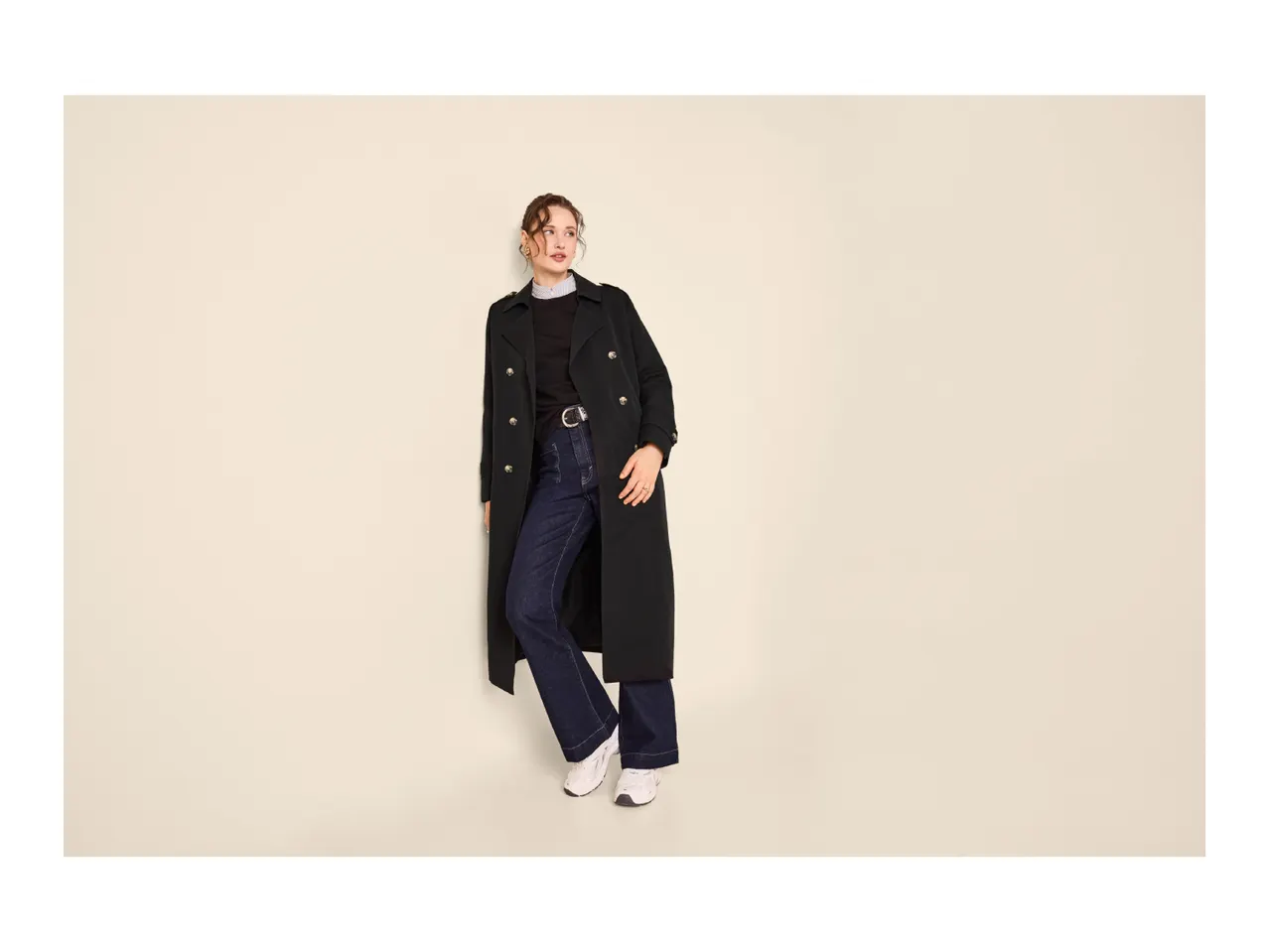 Woman in a black trench coat, dark jeans, and white sneakers, looking to the side.