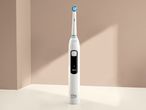 A white electric toothbrush with a blue and green brush head stands upright against a beige background.