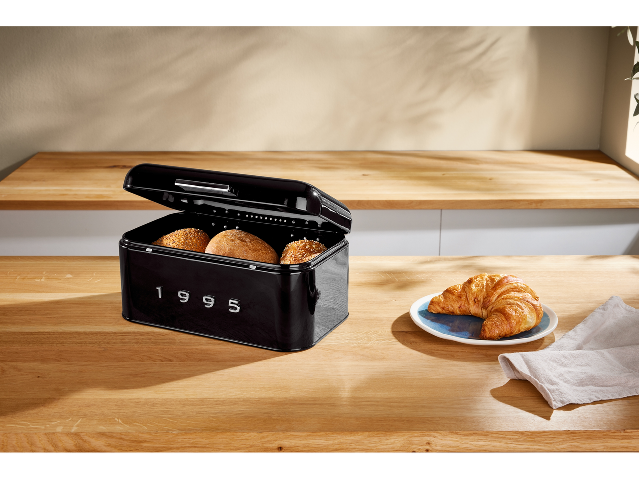 Black bread box with bread rolls and a croissant on a wooden counter.