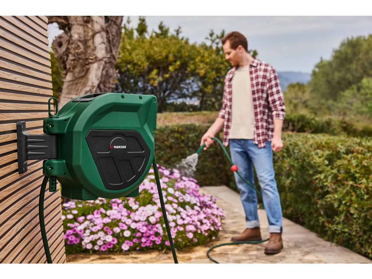 A man watering pink flowers with a green hose reel mounted on a wooden wall.