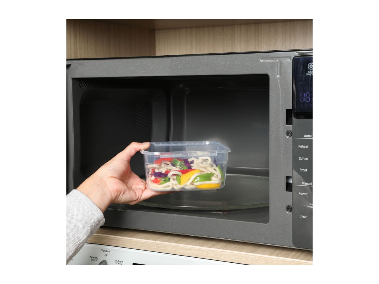 A hand placing a plastic food container with noodles and vegetables into a microwave