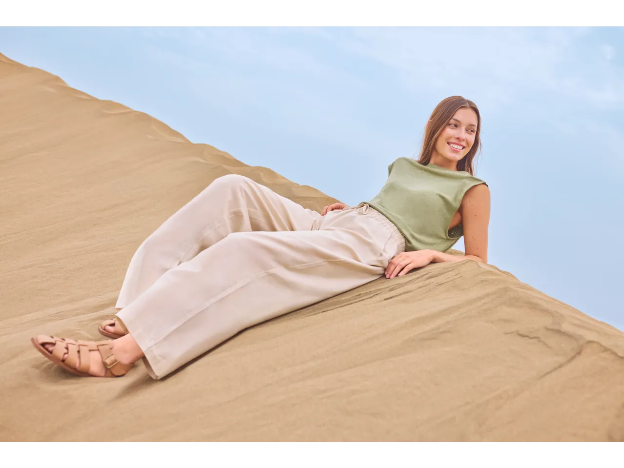 Woman in a green top and beige trousers lying on sand dunes, smiling.