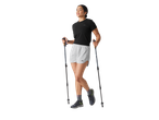 Mulher de short e camiseta preta, calçados esportivos e bastões de caminhada.