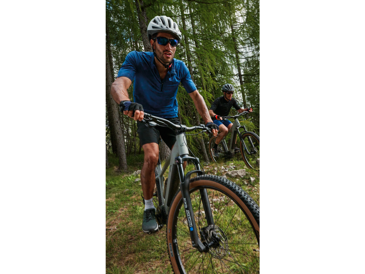 Two men mountain biking through a forest, wearing helmets, sunglasses, and athletic apparel.