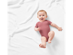 Un bebeluș zâmbitor într-un body roz, întins pe o pătură albă.