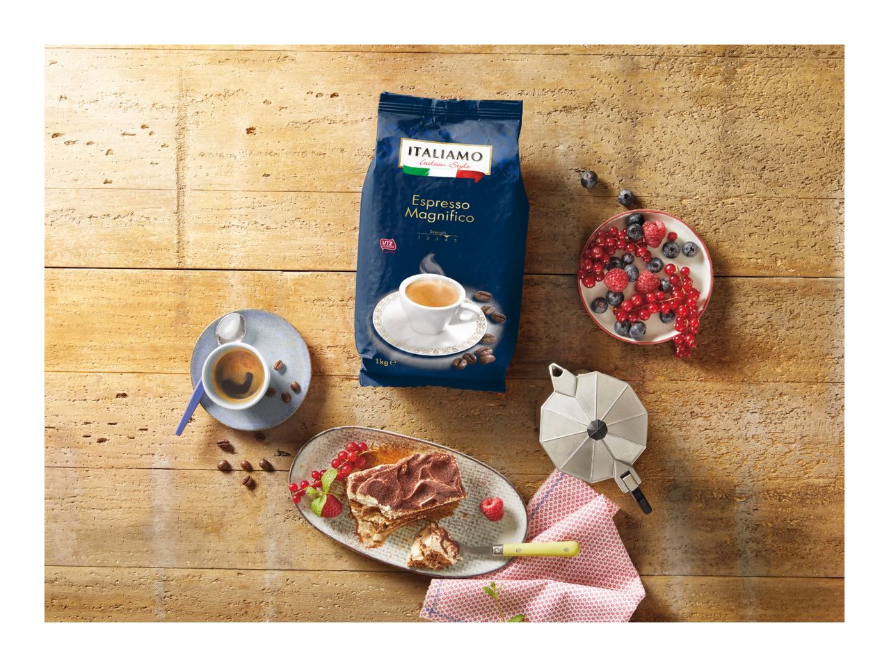 A bag of espresso coffee, a cup of coffee, tiramisu, berries, and a moka pot on a wooden table.