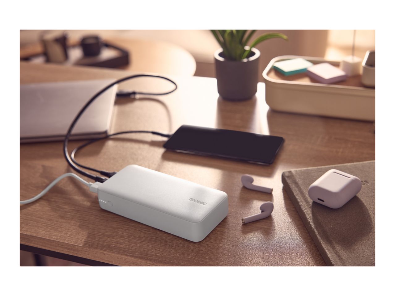 White power bank charging a smartphone and laptop on a wooden desk, with earbuds nearby.