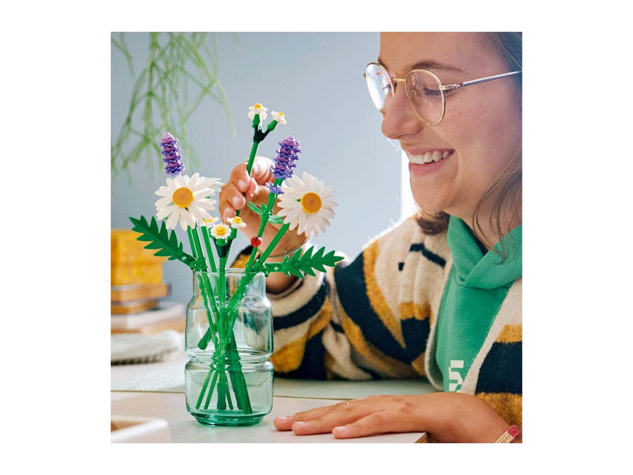 Smiling person arranging a bouquet of toy flowers in a glass vase.
