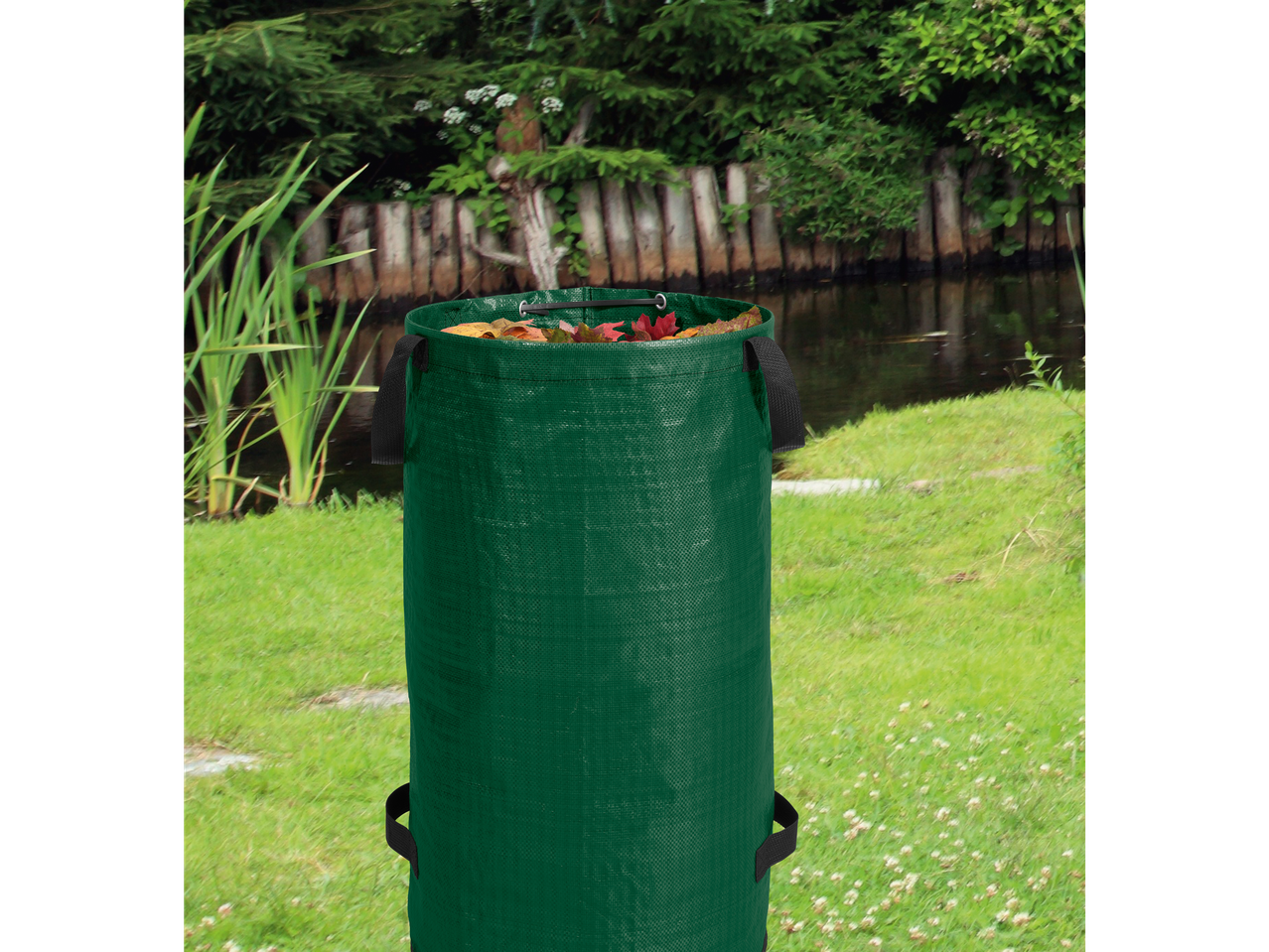 A green garden waste bag filled with autumn leaves sits on a grassy lawn near a pond.