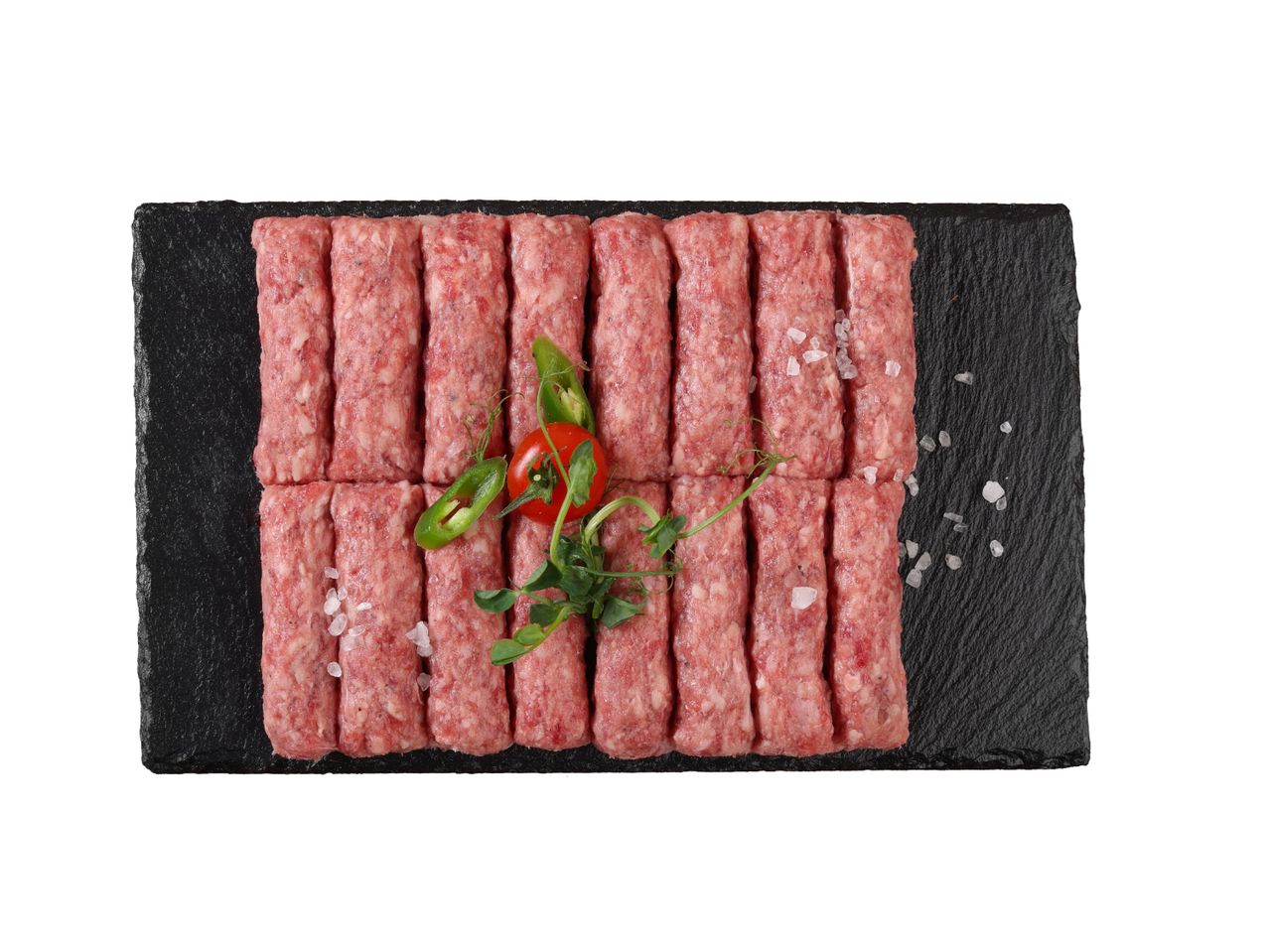 Sirove ćevapi poslagane na crnoj ploči, začinjeni solju i ukrašeni zelenilom.