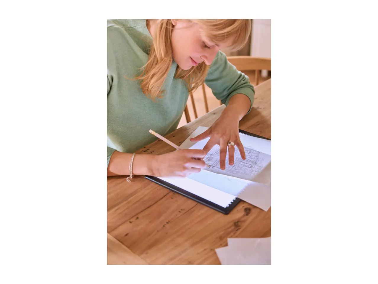 Woman tracing a drawing on a light pad with a pencil, on a wooden table.