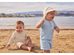 Two babies in muslin outfits on a sandy beach with the sea and mountains in the background.