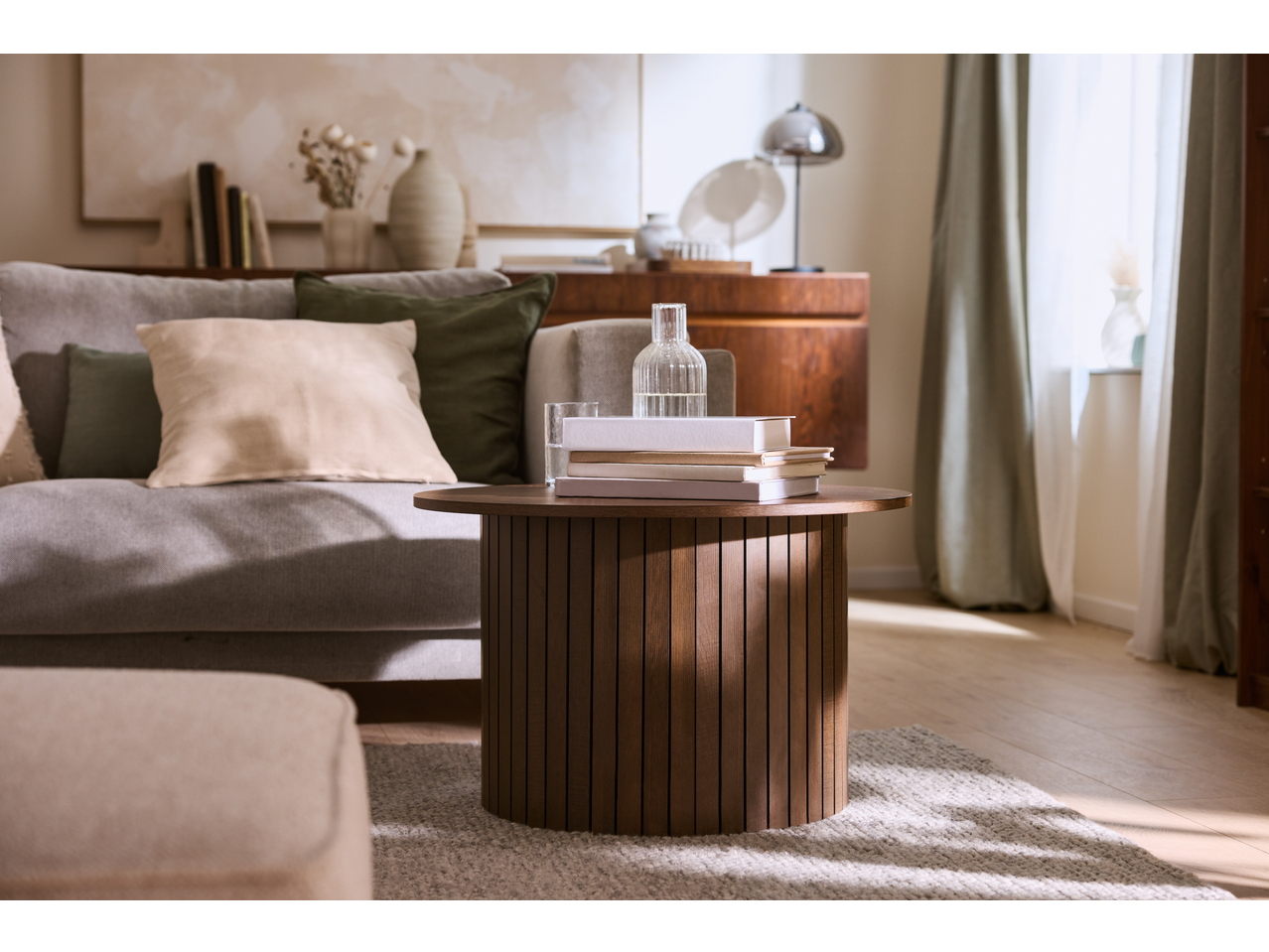 Living room with a round wooden coffee table, sofa with cushions, and a window with curtains.