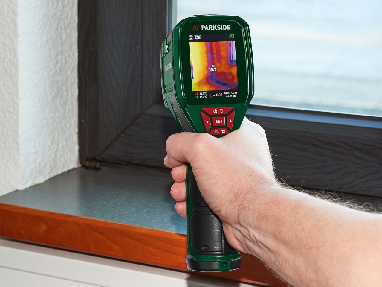 Hand holding a thermal imaging camera, displaying temperature readings near a window.