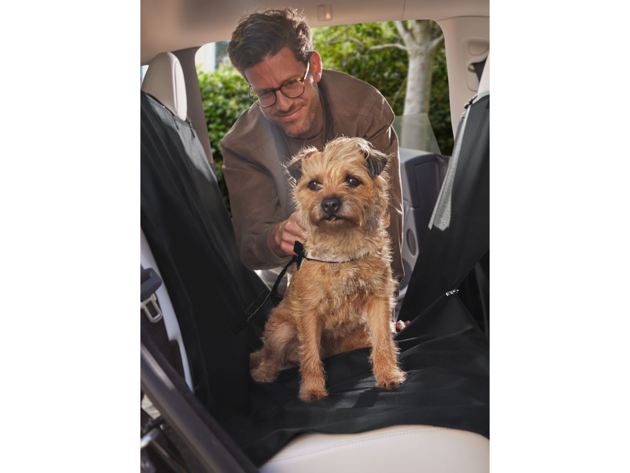 A man secures his dog in the backseat of a car using a pet car seat cover.