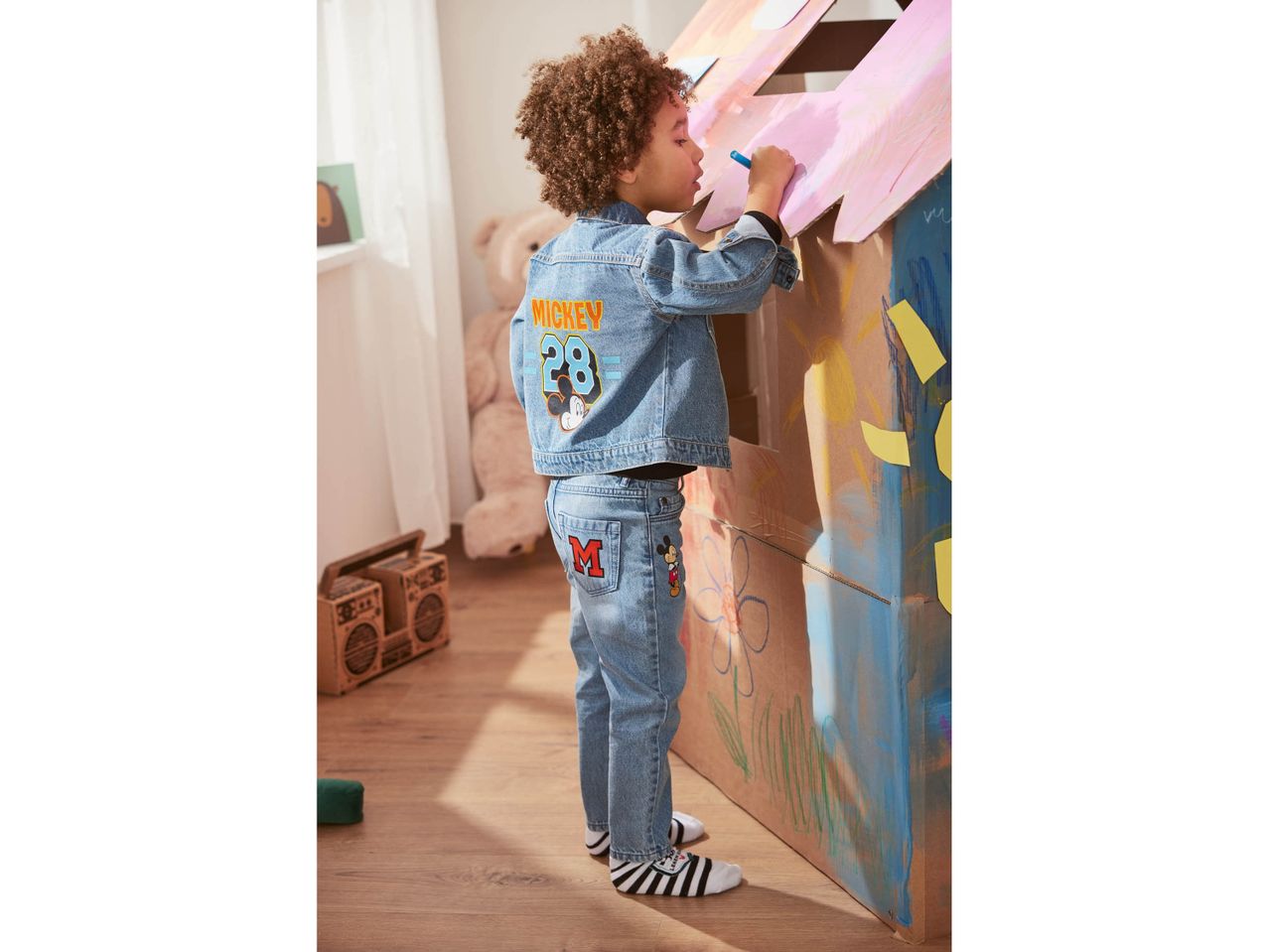 Child in Mickey Mouse denim jacket and jeans drawing on a cardboard playhouse.