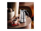 A stainless steel moka pot, a cup of coffee, and chocolates on a wooden table.