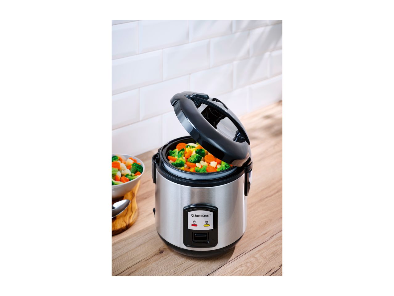 Electric rice cooker with steaming basket filled with vegetables on a wooden countertop.