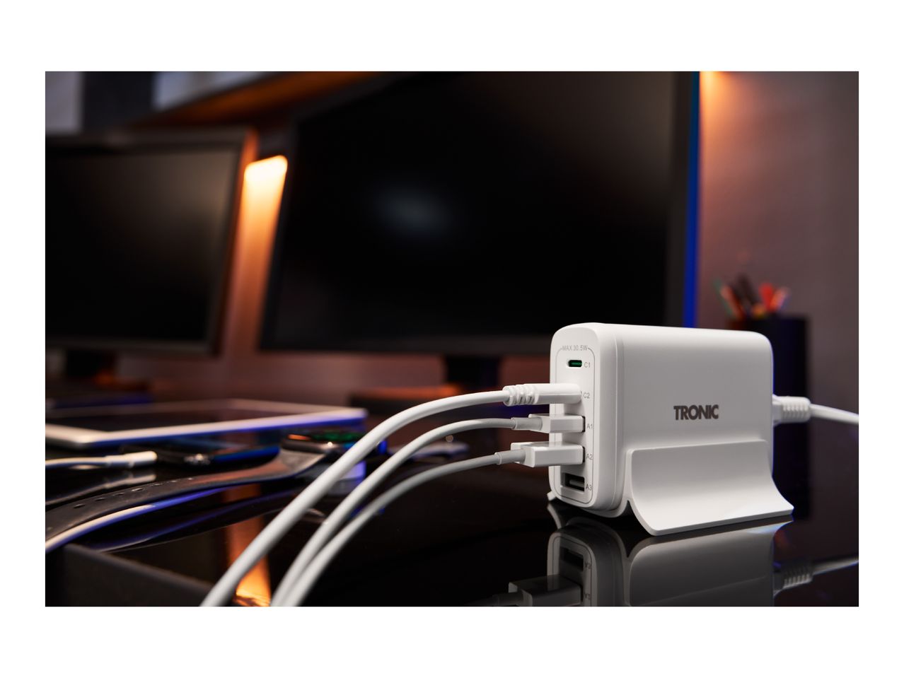 White multi-port charging station with several cables plugged in, on a desk with monitors.