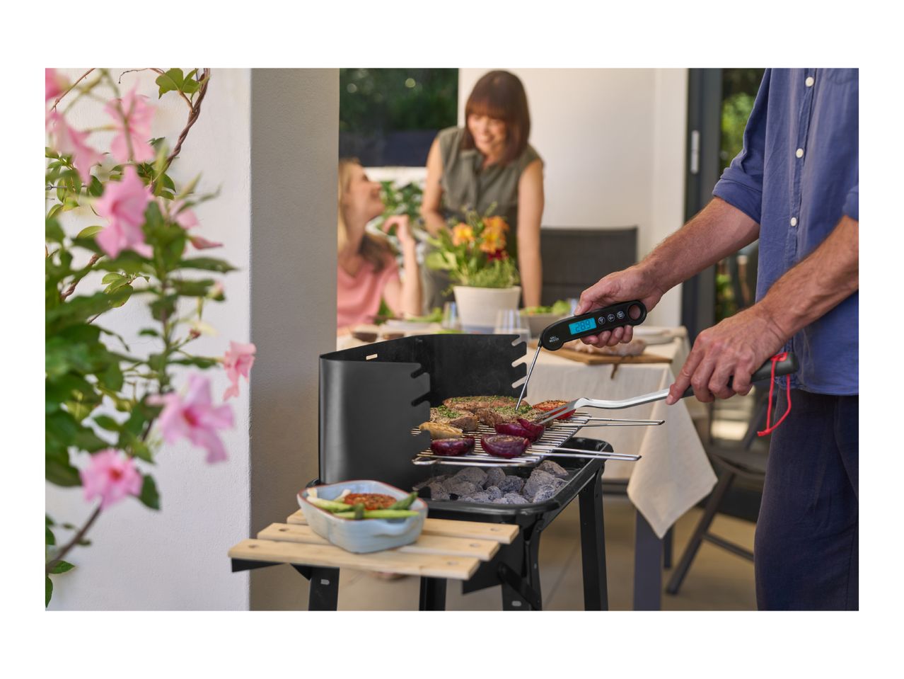 Man grilling meat and vegetables on a charcoal barbecue, using a digital thermometer and tongs.