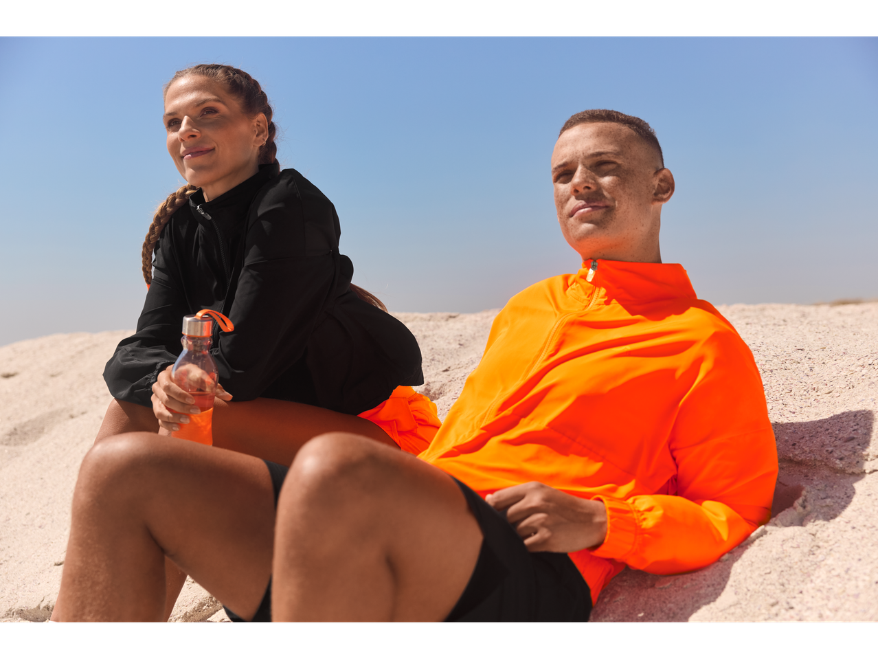 Doi atleți se odihnesc pe plajă după antrenament, unul cu o jachetă portocalie și celălalt cu una neagră.