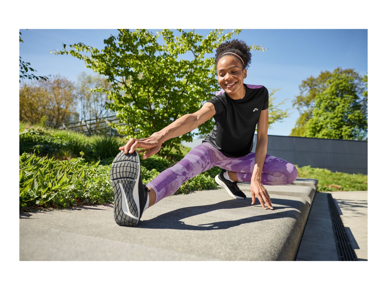 Woman in black and purple activewear stretching outdoors on a sunny day.