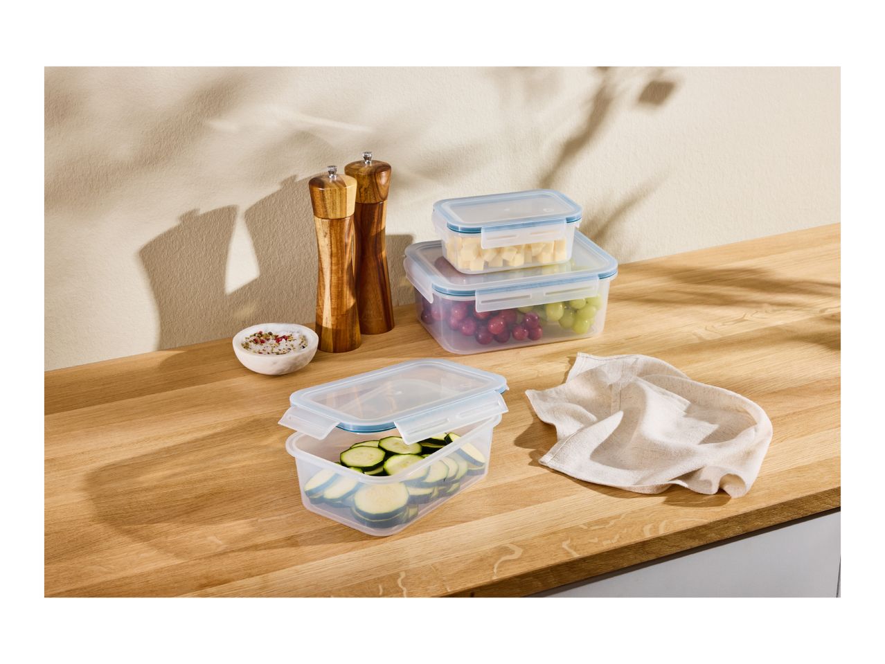 Food storage containers with grapes, cheese, and zucchini on a wooden counter with spices.