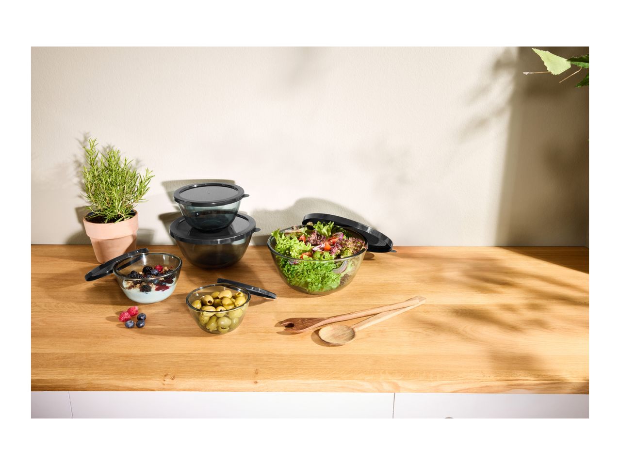 Glass bowls with dark lids containing salad, yogurt, and olives on a wooden counter.