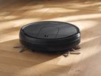 Black robotic vacuum cleaner on a wooden floor with sunlight and shadows.