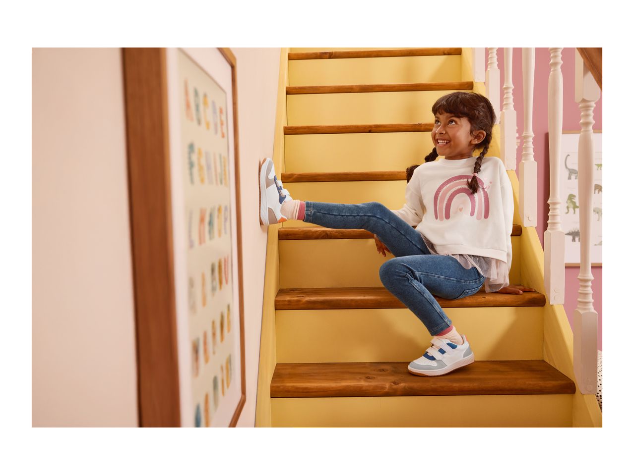 A child in a white rainbow sweatshirt and blue jeans sits on wooden stairs, wearing white and blue sneakers.