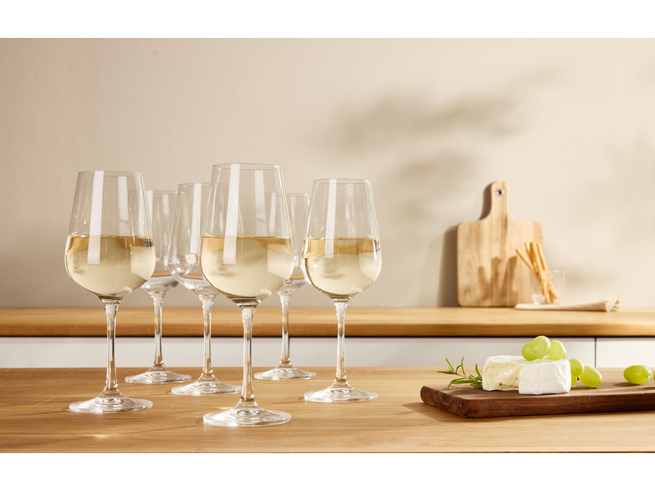 White wine glasses on a wooden counter with cheese, grapes, and breadsticks.
