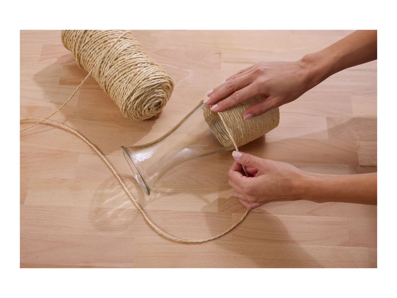 Hands wrapping a glass vase with natural twine on a wooden surface.