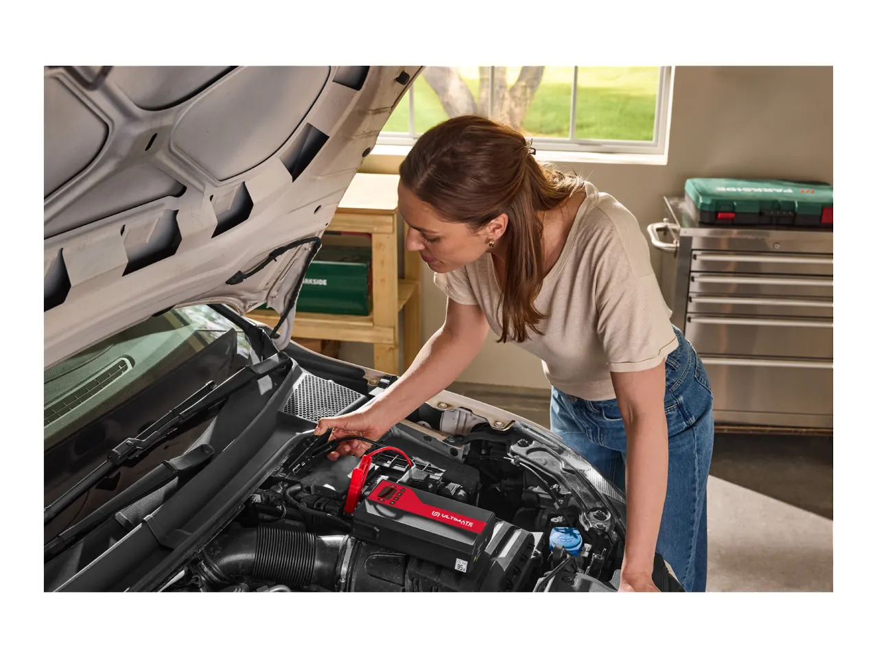 A woman uses a portable car battery charger under the open hood of a car.