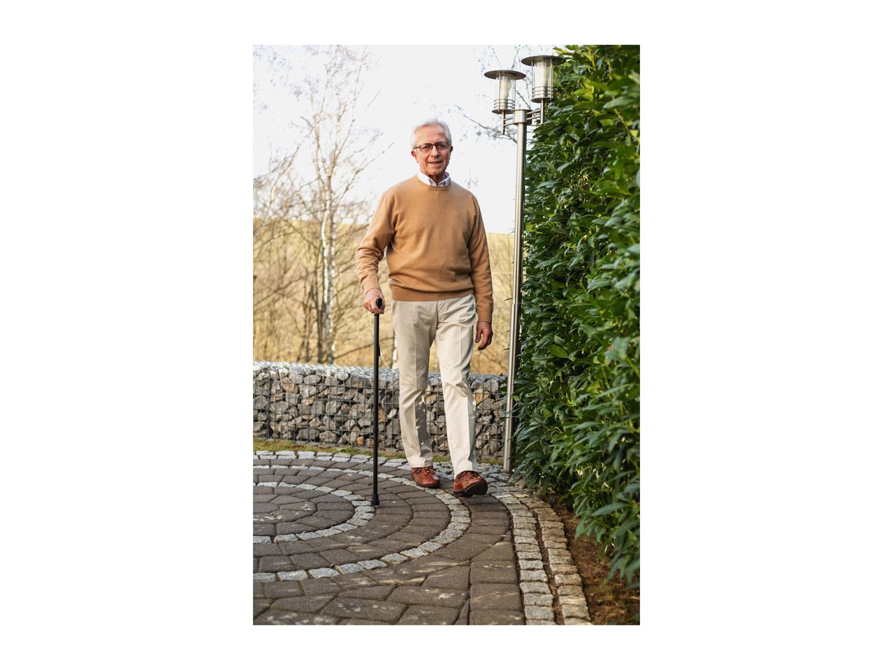 Elderly man walking with a cane on a paved path next to a green hedge.