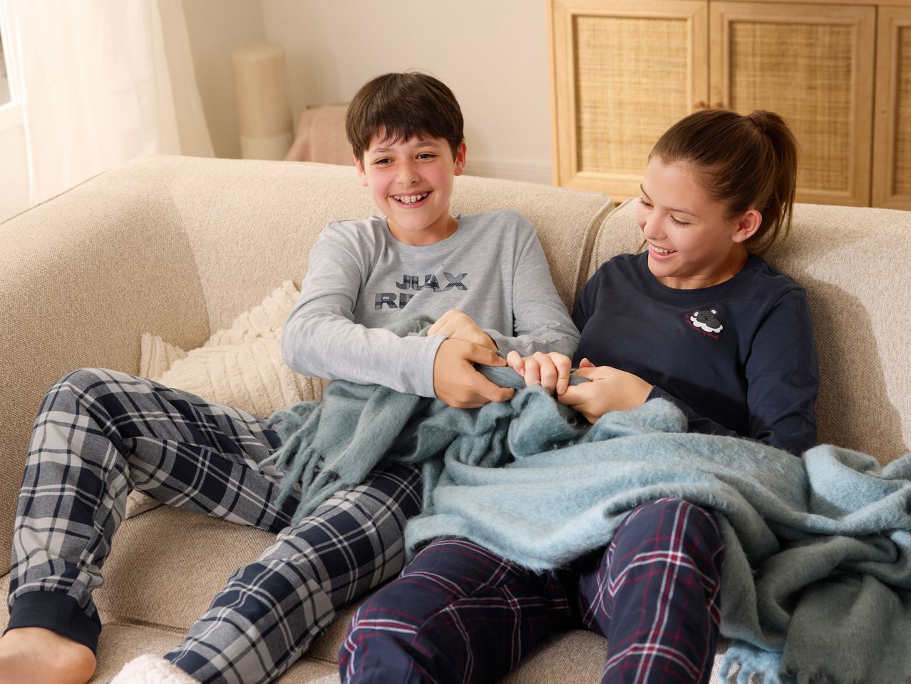 Two children in plaid pajamas and long-sleeved shirts sharing a blue blanket on a sofa.